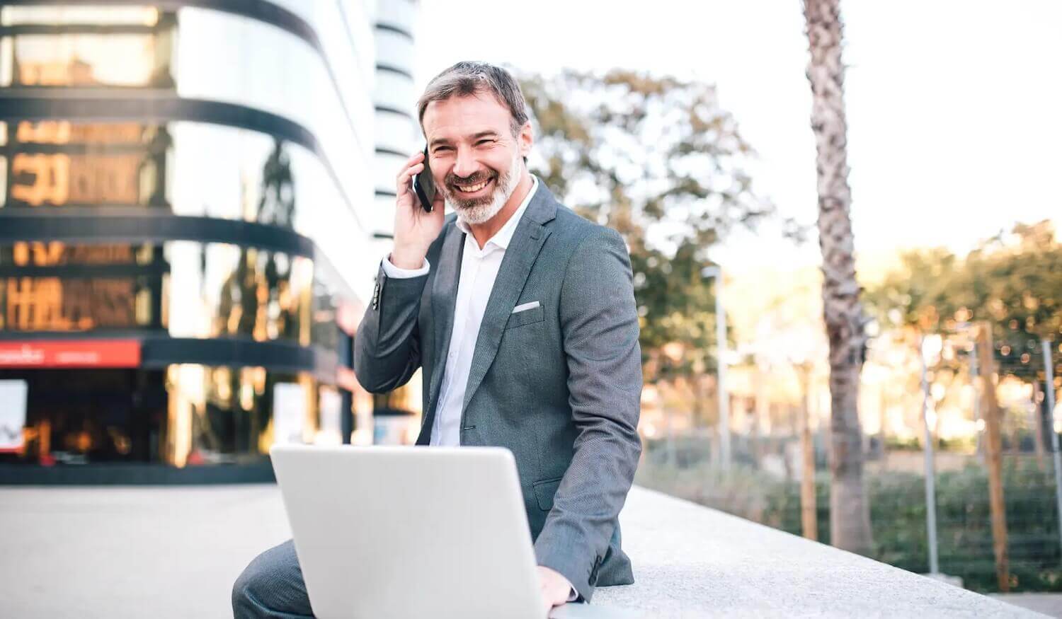 Financial services CEO sitting on a wall with laptop and phone outside office buildings