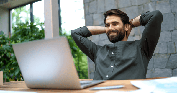 Professional man sitting at desk in front of laptop in office smiling with arms behind his head.png