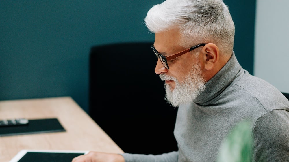 Older man sitting at desk with glasses on using laptop